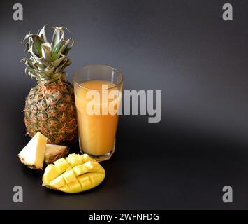 Un grand verre à facettes d'un mélange de jus de fruits sur fond noir, à côté de morceaux d'ananas mûr et de mangue. Gros plan. Banque D'Images