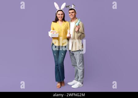 Heureux jeune couple avec panier de Pâques, lapins jouets et oreilles de lapin sur fond lilas Banque D'Images