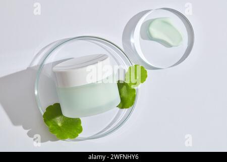Un pot d'étiquette vide placé sur un podium transparent en verre avec des feuilles de gotu kola et une texture crème. Maquette de produit d'extrait de Gotu kola (Centella asiatica) Banque D'Images