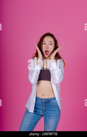 Jeune femme choquée avec les mains encadrant le visage et la bouche ouverts dans la surprise, sur fond rose. Banque D'Images