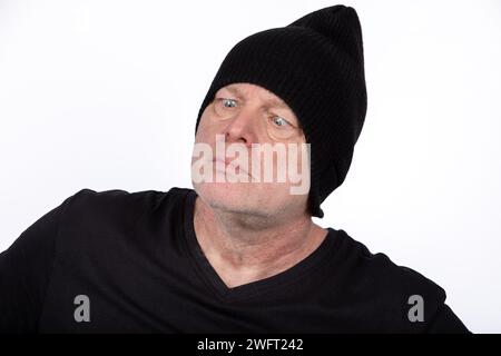 Homme d'âge moyen décalé en T-shirt noir et chapeau avec expression croisée, personnage humoristique sur fond blanc - concept de portrait excentrique Banque D'Images