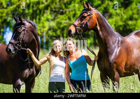 La photo montre deux jeunes femmes en vêtements légers d'équitation d'été avec de longs cheveux bruns, debout avec leurs chevaux sur une prairie d'été et riant moi Banque D'Images