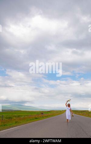 Une fille en robe blanche marche avec des fleurs sur la route Banque D'Images