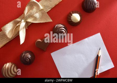 Concept de cadeau d'amour avec bonbons au chocolat sur fond rouge avec noeud doré et papier vierge avec crayon. Vue de dessus. Banque D'Images
