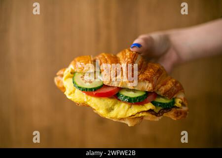 un croissant fourré de légumes concombre tomate et oeufs frits omelette sur la main femelle sur un fond en bois, vue de dessus Banque D'Images