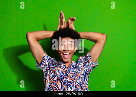 Photo de souriant heureux gars fou plaisantant les oreilles des paumes ressemble à un lapin ont du plaisir à porter une chemise à la mode isolé sur fond de couleur verte Banque D'Images
