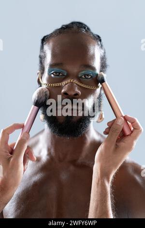Portrait de l'homme gay afro-américain appliquant le maquillage avec des pinceaux tenus par le crop artist. Gros plan d'un homme homosexuel portant un fard à paupières lumineux et une chaîne d'or regardant la caméra sur fond bleu. Banque D'Images