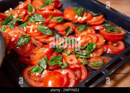 Tranches de tomates assaisonnées de basilic frais, d'ail et de poivre sur une plaque de cuisson, préparées pour rôtir. Banque D'Images