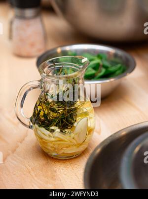 Une cruche en verre remplie d'huile d'olive infusée d'herbes fraîches, d'ail et de tranches de citron, posée sur un comptoir de cuisine en bois. Banque D'Images