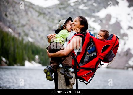 Une mère porte son fils de 6 mois dans un bébé sac à dos de randonnée, tandis qu'elle tient et embrasse son autre fils. Banque D'Images