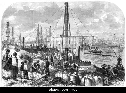 Le déluge dans les fens : faire le cofferdam, 1862. 'Les espoirs, mêlés en grande partie à des craintes, des agriculteurs des marais sont centrés sur le gigantesque cofferdam que M. Leather, l'entrepreneur, a poussé à travers le drain de Middle Level, à environ 800 mètres au-dessus de l'écluse explosée. La charpente de ce barrage se compose d'un grand nombre de pieux... qui sont enfoncés à une profondeur de vingt pieds dans la terre. Tous ces pieux sont entourés ensemble par des boulons de fer fixés dans le lit de la rivière par des plongeurs experts... les rives étaient tapissées de sacs d'argile au nombre de plusieurs milliers, et les corps des hommes étaient enga Banque D'Images