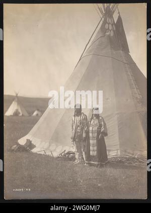 AIMS Back, 1910. La photographie montre AIMS Back avec femme, à la fois en robe traditionnelle, debout à l'extérieur du tipi. Banque D'Images