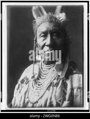 Spectacles de poissons, un Indien Apsaroke, portrait tête et épaules, face à face, pompadour, tresses de temple peintes en blanc, 2 plumes d'aigle, boucle d'oreille disque, perles de coquille avec de grands ornements de disque, chemise en peau de mousseline perlée, c1908. Banque D'Images