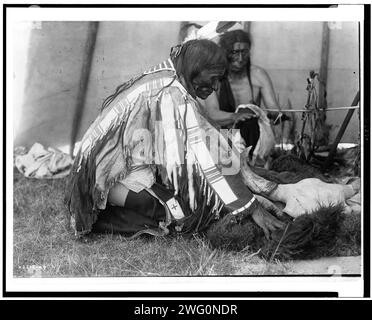 Hu Kalowa Pi-enlève le revêtement, c1907. Intérieur du tipi, homme agenouillé sur le sol remouillant la peau de buffle autour du crâne sur le sol, un autre mâle derrière l'autel réchauffant les mains par le feu. Banque D'Images