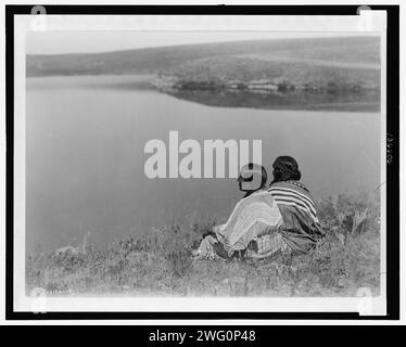 Une heure d'inactivité, Piegan, c1910. La photographie montre deux Indiens Piegan assis sur une zone herbeuse au-dessus d'un plan d'eau. Banque D'Images