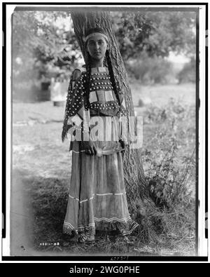 Jeune fille Sioux, c1908. Portrait, pleine longueur, debout près de l'arbre, serre-tête perlé, robe en peau de mousqueton perlée. Banque D'Images