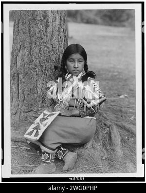 Umatilla enfant, c1910. Fille Umatilla, portrait en pied, face à l'avant, assise au sol près du pin, portant une robe de couverture perlée, des mocassins perlés, tenant le sac perlé sur les genoux. Banque D'Images