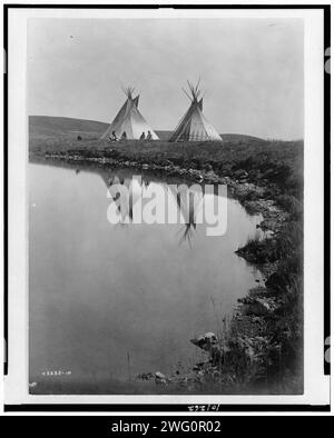 Au bord de l'eau-Piegan, c1910. La photo montre deux tipis reflétés dans l'eau de l'étang, avec quatre Indiens Piegan assis devant un tipi. Banque D'Images