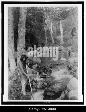 Le ruisseau à saumon, c1923. La photographie montre un homme tenant une lance tout en se tenant debout sur des rochers près d'un ruisseau courant. Banque D'Images