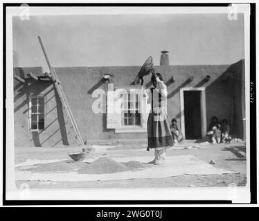 Vendange du blé, c1905. Femme Tewa remuant du blé devant le bâtiment pueblo. Banque D'Images