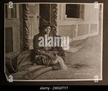 Femme maorie préparant le lin. 1891. Une femme maorie avec un enfant est assise près de l ' entrée d ' une maison maorie en bois. Elle a les cheveux courts, des boucles d'oreilles et une pipe dans la bouche. Elle porte une robe européenne. Elle tord des fibres de lin sur sa jambe pour les préparer au tissage. Portrait en pied. Du voyage de Charles Appleton Longfellow en Nouvelle-Zélande en 1891. Banque D'Images