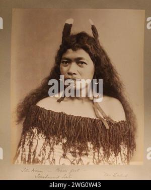 Une fille maorie, nom de famille Titia incertain, pose sur un fond clair. Elle a des cheveux longs aux épaules décorés de plumes et de rubans et des tatouages faciaux autour de sa bouche. Elle porte un manteau rigide, décoré de cordes pendantes, tiré vers le bas autour de ses épaules. Portrait de la tête et des épaules. Du voyage de Charles Appleton Longfellow en Nouvelle-Zélande en 1891. Banque D'Images