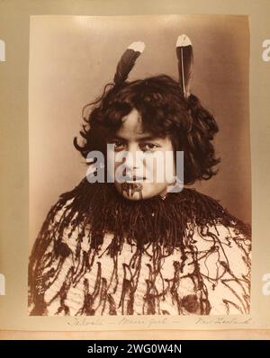 Une fille maorie, Tekoata, pose sur un fond ordinaire. Elle a des cheveux de longueur moyenne décorés de plumes et de tatouages faciaux autour de sa bouche. Elle porte un manteau rigide, décoré de cordes pendantes, autour de ses épaules. Portrait de la tête et des épaules. Du voyage de Charles Appleton Longfellow en Nouvelle-Zélande en 1891. Banque D'Images