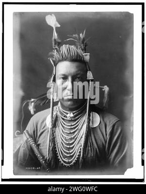 Swallow Bird, Crow Indian, Montana, portrait tête et épaules, face avant, pompadour peint, coiffe Apsaroke, chemise en flanelle, perles sur la poitrine, pipe cloutée tenue devant le bras droit, c1908. Banque D'Images