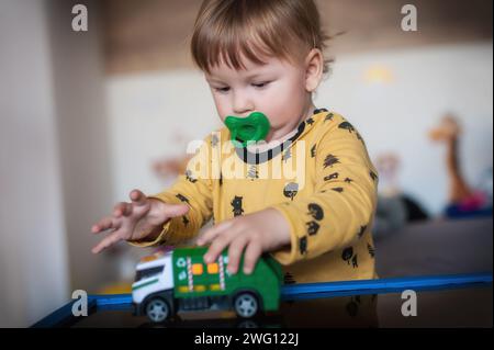 Mignon bébé garçon avec sucette dans la bouche joue avec un petit camion jouet, le conduisant autour d'un monde de fait-croire rempli d'excitation et d'émerveillement Banque D'Images