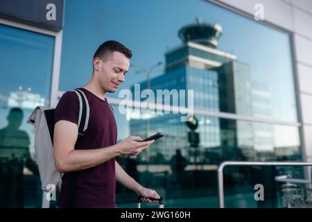 Voyageur commandant un taxi via une application mobile. Accent sélectif sur l'homme souriant avec sac à dos regardant le téléphone intelligent contre le terminal de l'aéroport sur l'été ensoleillé da Banque D'Images