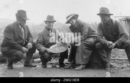 Portrait de groupe d'une femme et de trois hommes, accroupis dehors, souriant ; l'un des hommes expose un plat d'or bronzé, entre c1900 et 1916. Banque D'Images