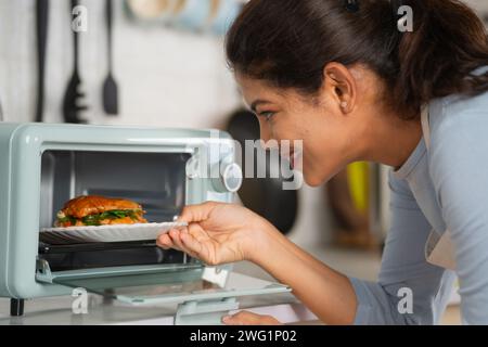 Heureuse femme indienne cuisinant un hamburger en le plaçant sur un four à micro-ondes à la cuisine - concept d'une alimentation saine, d'un mode de vie domestique et de la technologie Banque D'Images