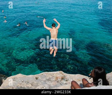 Un homme saute des rochers dans la mer, tandis qu'une femme fume, sur une plage privée à Valetta, Malte en été. Banque D'Images