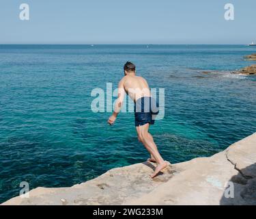 Un homme saute dans la mer des rochers sur une plage privée à Valetta, Malte en été. Banque D'Images