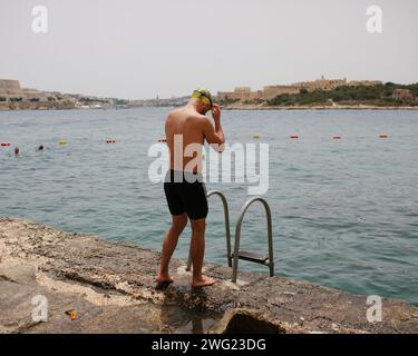 Un nageur se prépare à entrer dans la mer à la plage de Tigne point, Malte. La Valette peut être vue en arrière-plan. Banque D'Images