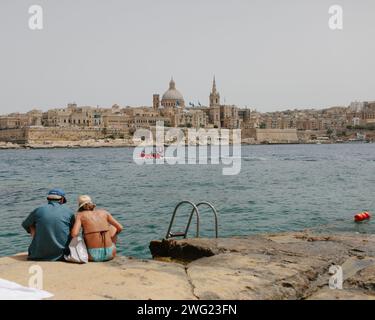 Un couple âgé s'assoit sur les rochers donnant sur la mer vers le front de mer de la Valette à Malte un jour d'été. Banque D'Images