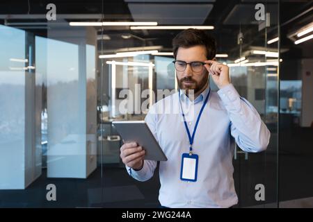 Confus et sérieux jeune homme spécialiste et développeur est dans les locaux du centre de bureaux, debout dans une chemise et avec un badge, participant à une conférence d'ingénierie, tenant des lunettes et utilisant une tablette. Banque D'Images