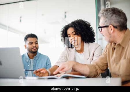 Une femme d'affaires concentrée en blazer chic se penche pour discuter d'un document avec ses collègues, démontrant le travail d'équipe et la résolution active de problèmes dans un bureau contemporain. Équipe de travail résolvant les tâches ensemble Banque D'Images