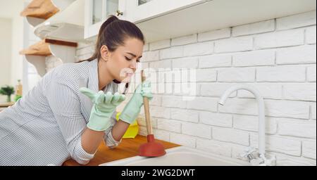 Jeune femme brune nettoyant évier bloqué et drain avec un piston dans la cuisine à la maison. Banque D'Images