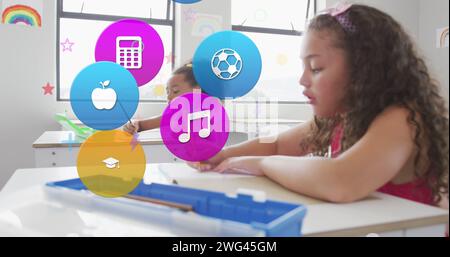 Image des icônes de matière scolaire sur les écolières heureuses et diverses écrivant dans la classe Banque D'Images