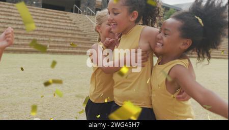 Image de confettis dorés sur des écolières joyeuses diverses célébrant et serrant sur le terrain de sport Banque D'Images