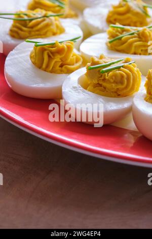 Gros plan d'œufs farcis avec deux brins de ciboulette sur une assiette avec un bord rouge sur une table en bois. Banque D'Images