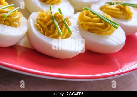 Gros plan d'œufs farcis avec deux brins de ciboulette sur une assiette avec un bord rouge sur une table en bois. Banque D'Images