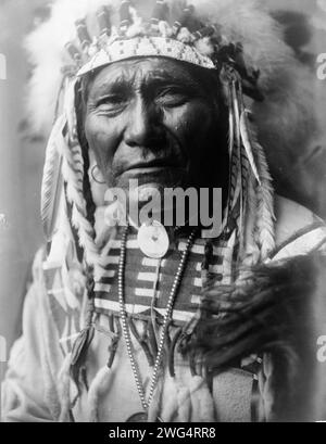 Ghost Bear, Crow Indian, Montana, portrait tête et épaules, face avant, coiffe en plumes, chemise en peau de busard perlée, collier de perles avec concho en coquille, grande médaille sur brin de perles métalliques, c1908. Banque D'Images