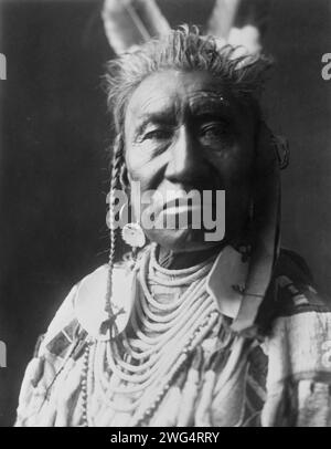 Spectacles de poissons, un Indien Apsaroke, portrait tête et épaules, face à face, pompadour, tresses de temple peintes en blanc, 2 plumes d'aigle, boucle d'oreille disque, perles de coquille avec de grands ornements de disque, chemise en peau de mousseline perlée, c1908. Banque D'Images