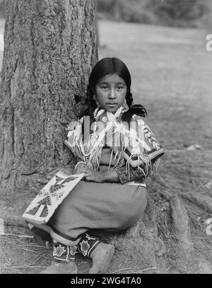 Umatilla enfant, c1910. Fille Umatilla, portrait en pied, face à l'avant, assise au sol près du pin, portant une robe de couverture perlée, des mocassins perlés, tenant le sac perlé sur les genoux. Banque D'Images