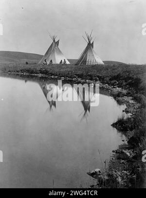 Au bord de l'eau-Piegan, c1910. La photo montre deux tipis reflétés dans l'eau de l'étang, avec quatre Indiens Piegan assis devant un tipi. Banque D'Images