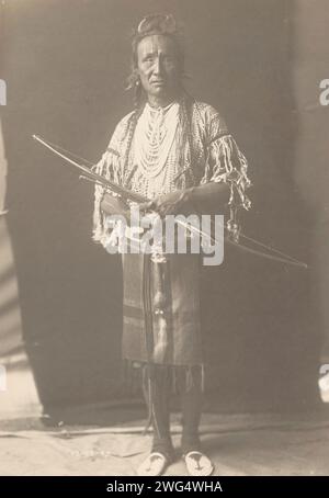 Crow Chief, 1910. La photographie montre le portrait en pied de l'ours grizzli, avec les cheveux tressés, portant des vêtements traidtional et plusieurs colliers, debout, tenant l'arc, face à l'avant. Banque D'Images