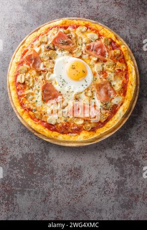 Pizza italienne Bismarck garnie de sauce tomate, fromage fondu, oeuf, champignons, jambon gros plan sur une planche de bois sur la table. Vue verticale de dessus depuis ab Banque D'Images