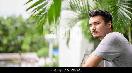 Jeune homme en chemise grise appuyé sur une balustrade, perdu dans ses pensées, avec des plantes tropicales Banque D'Images